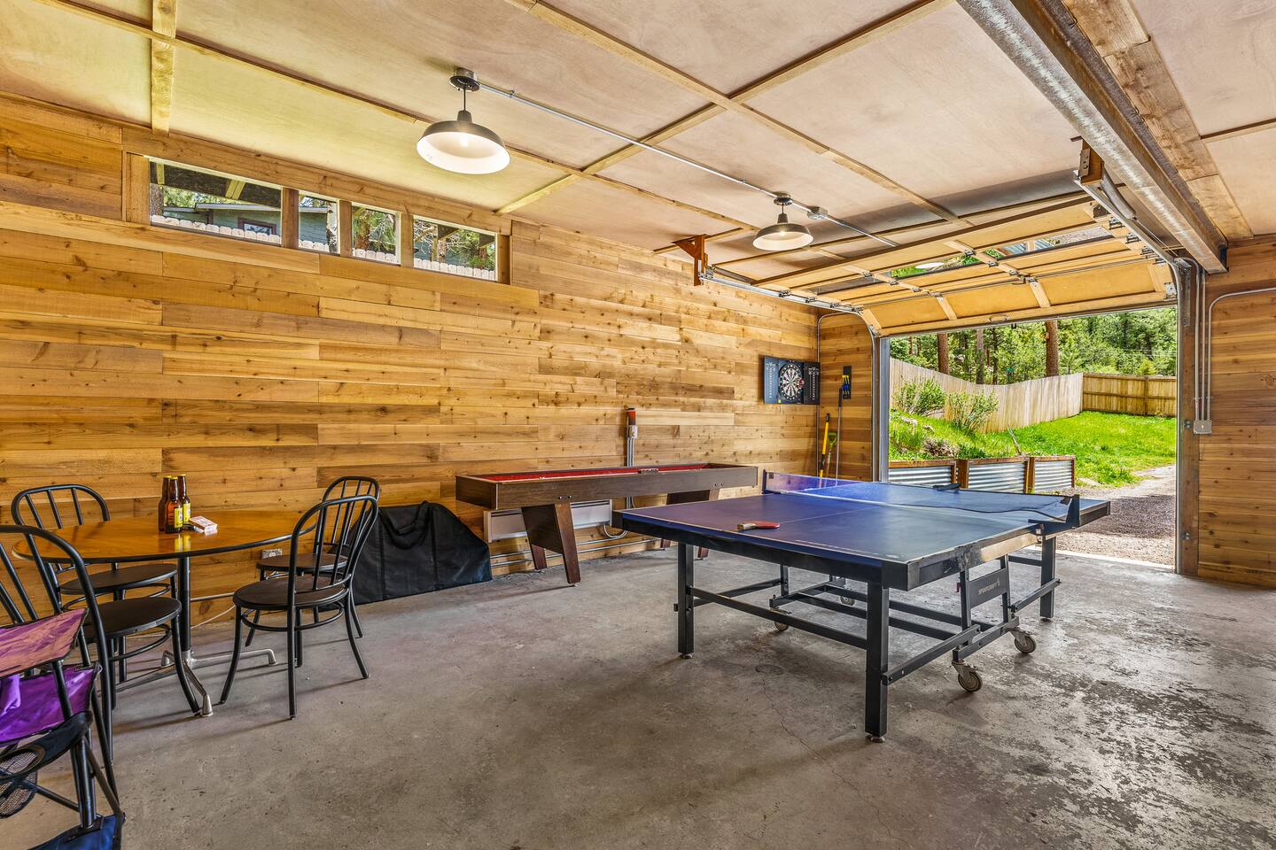 Game room with a ping pong table, a card table and a wood-paneled wall, with the garage door open to the trees