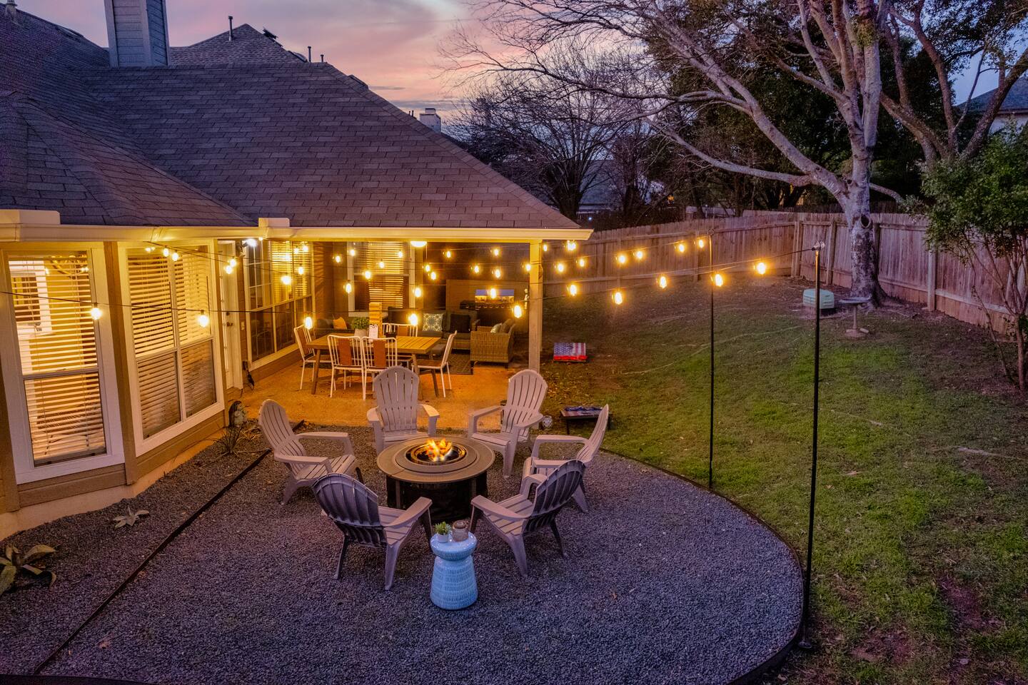 Twilight Patio Retreat! ✨ Enjoy the gorgeous sunset from the string-lit covered patio featuring an outdoor dining table. Gather around the fire pit with  Adirondack chairs set on a pebble courtyard. Fully fenced and perfect for evening relaxation!