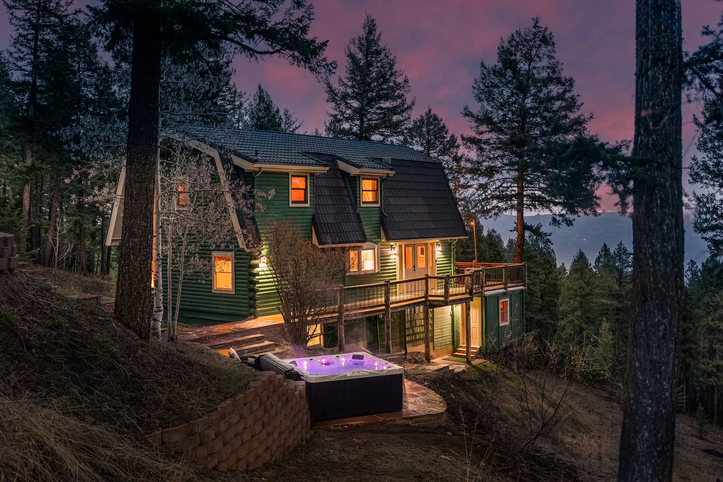 The green barn-style home lit up at dusk among the pines