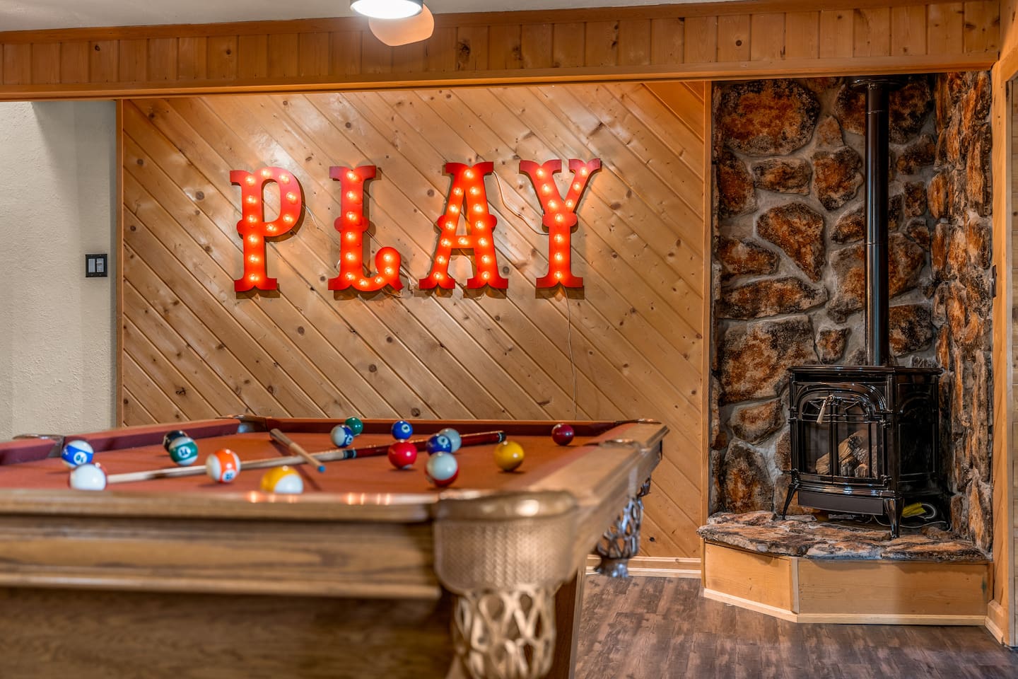 Pool table under a lit PLAY marquee sign beside a stone wood stove