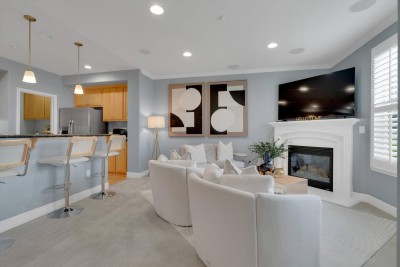 Chic Open-Concept Living! ✨️ Relax in this light-filled living room featuring a cozy fireplace, large Smart TV, and plush white seating. The space flows into the kitchen with bar seating for three. Stylish art complete this perfect gathering spot.