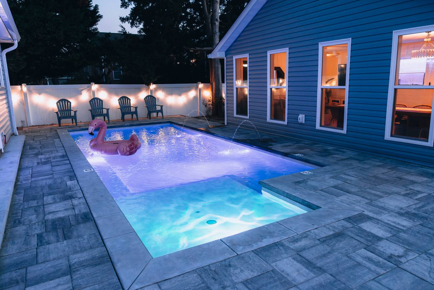 Pool with fountain jets and four Adirondack chairs, house windows glowing