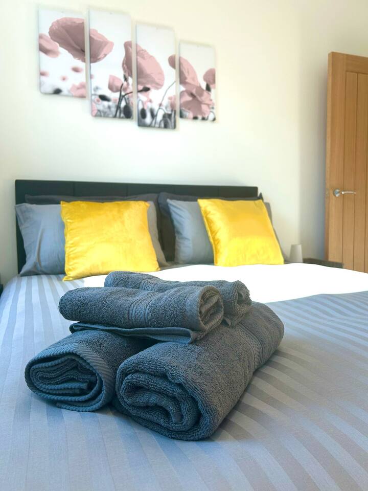 Simple and cozy bedroom on Wilton Road, Greater London, near Notting Hill, Acton, and Ealing. Features a double bed with yellow pillows, grey curtains, wooden flooring, a yellow armchair, and pretty floral wall art.