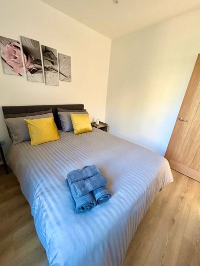 Bright bedroom on Wilton Road, Greater London, near Notting Hill, Acton, and Ealing. The space has a cozy double bed with yellow pillows, grey curtains, wooden floors, a bright yellow armchair, and floral wall art.