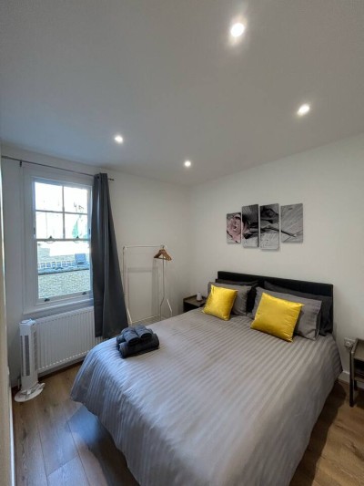 Stay at Wilton Road, Greater London, near Notting Hill, Acton, and Ealing. This bedroom features a cozy double bed with yellow accent pillows, stylish grey curtains, wooden flooring, a yellow armchair, and floral wall art.