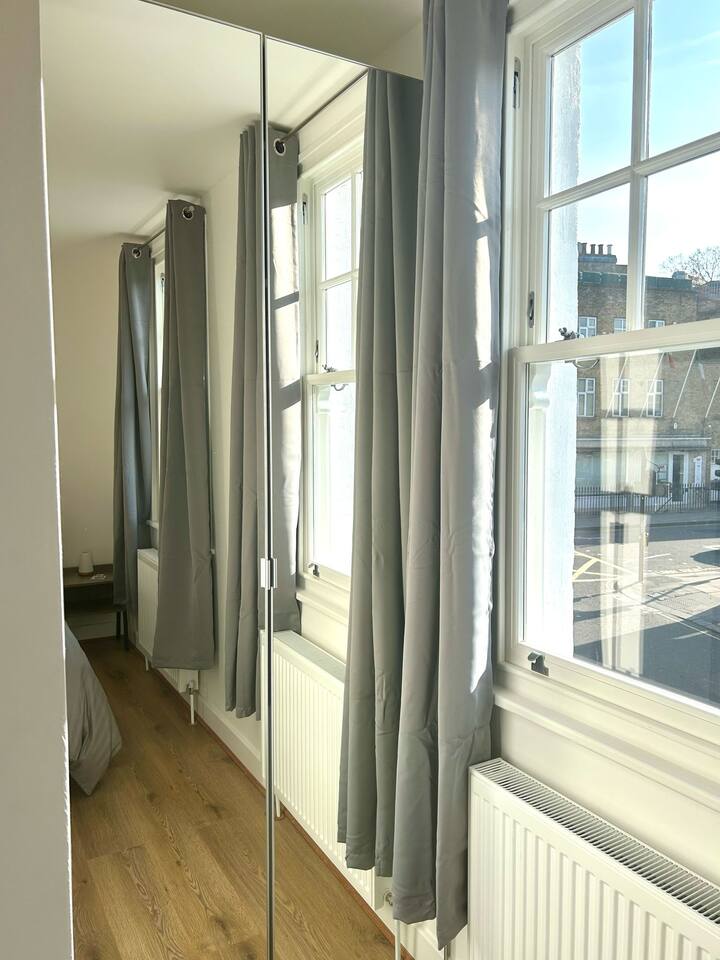 Bedroom on Wilton Road, Greater London, close to Notting Hill, Acton, and Ealing. Includes a cozy double bed with yellow accent pillows, stylish grey curtains, wooden floors, a bright yellow armchair, and floral wall art