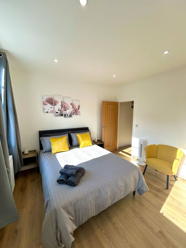 Bedroom on Wilton Road, Greater London, in the Notting Hill, Acton, and Ealing area. Comes with a cozy double bed, yellow pillows, grey curtains, wooden floors, a yellow armchair, and floral wall art. Perfect for a simple and comfortable stay.