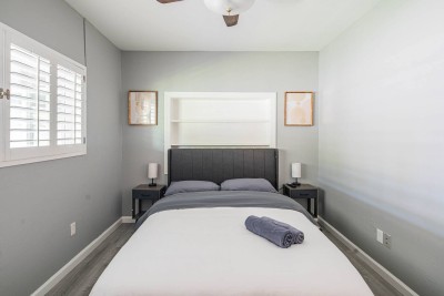 This relaxing space features a comfortable bed with a sleek upholstered headboard. The walls are painted a calming gray, and the window has classic plantation shutters. Includes built-in shelving and two dedicated nightstands with lamps. 