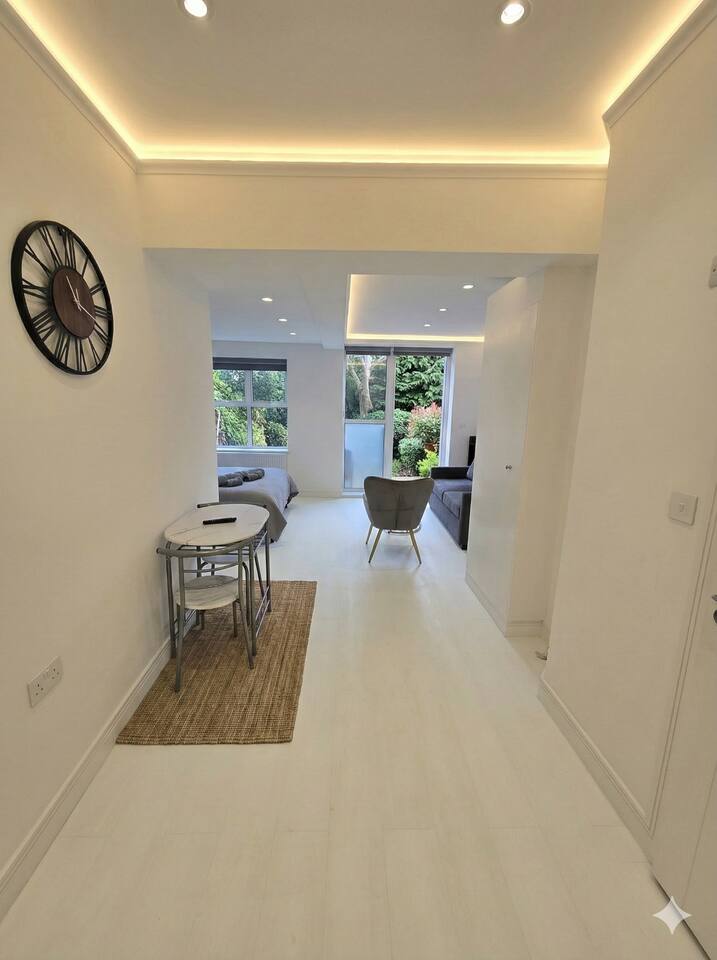 Modern, sun-drenched studio with a plush bed, dining area, and leafy garden views.