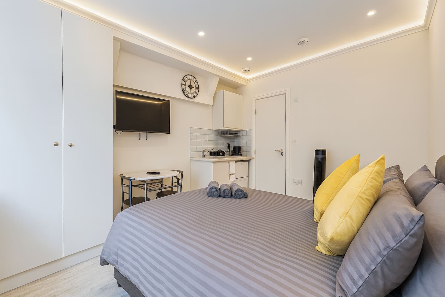 Cozy studio with grey bedding, kitchenette, TV, and warm modern lighting.