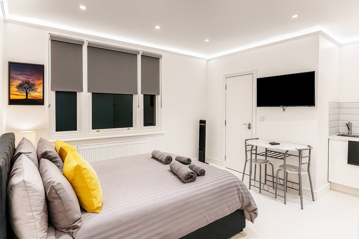 Bright studio with grey and yellow accents, TV, kitchenette, and large windows.