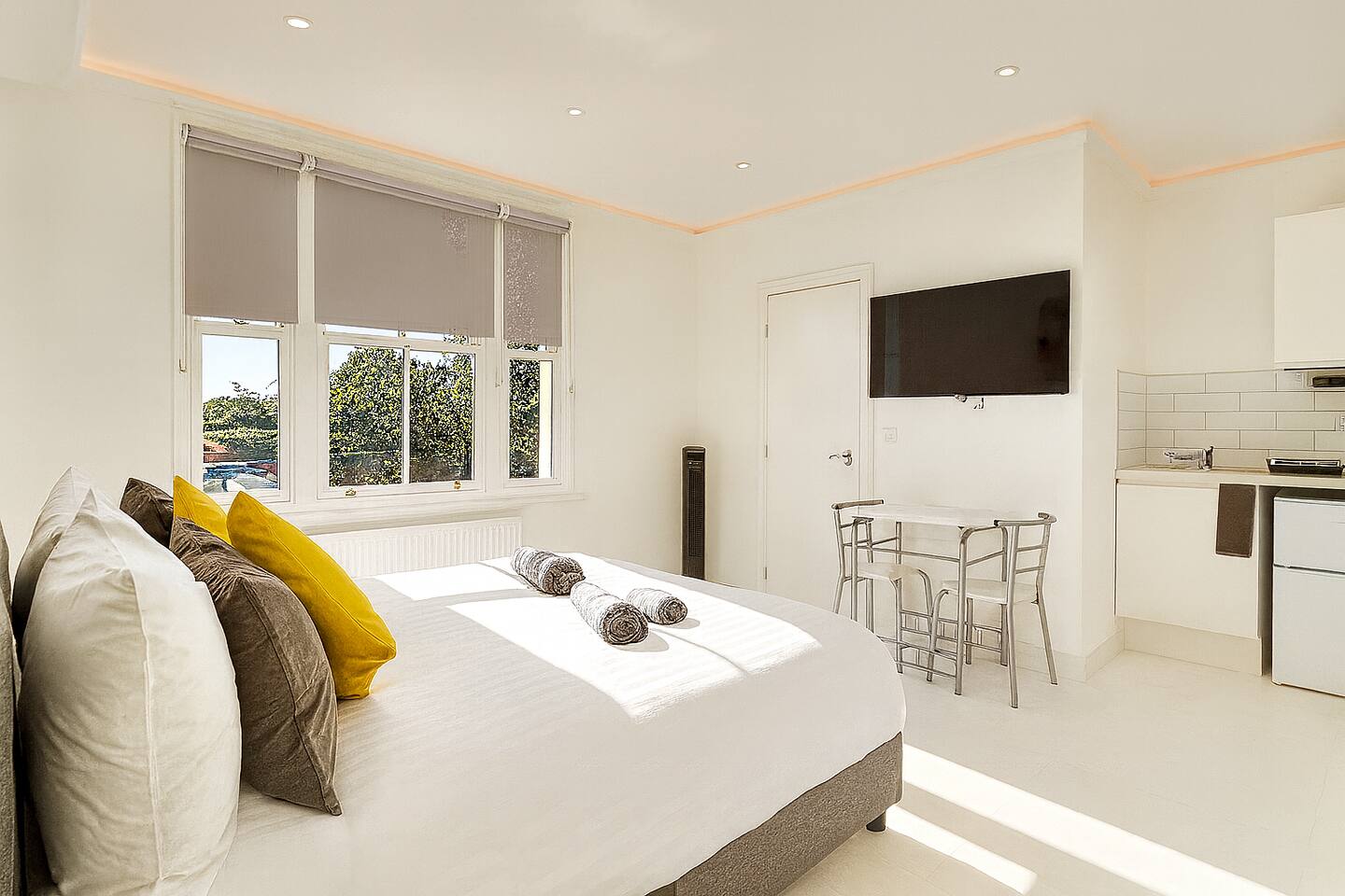 Bright studio with white bedding, yellow accents, kitchenette, and large window view.