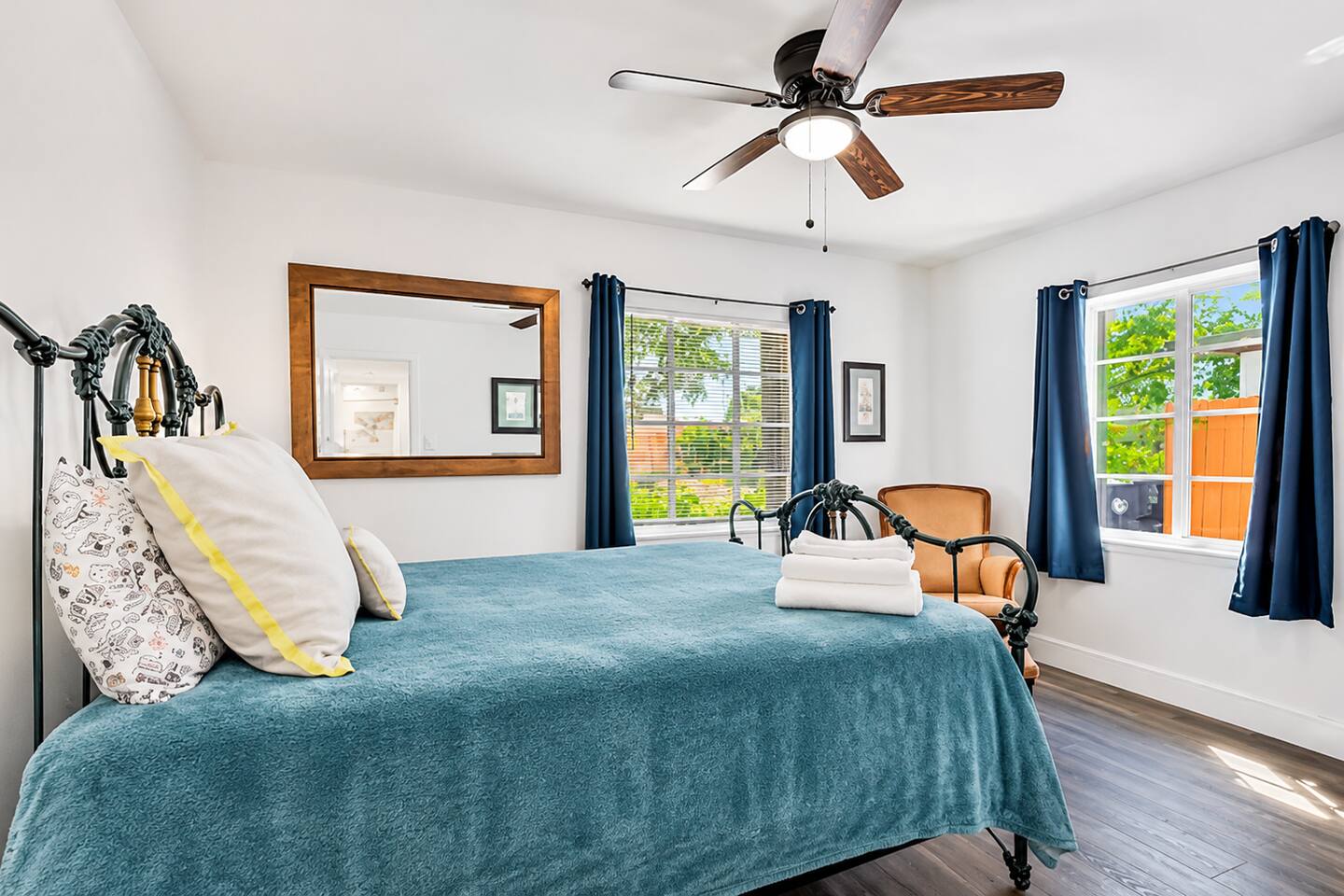 Inviting bedroom with teal metal bed, folded towels & garden views — includes cushioned armchair, ceiling fan & cozy natural light