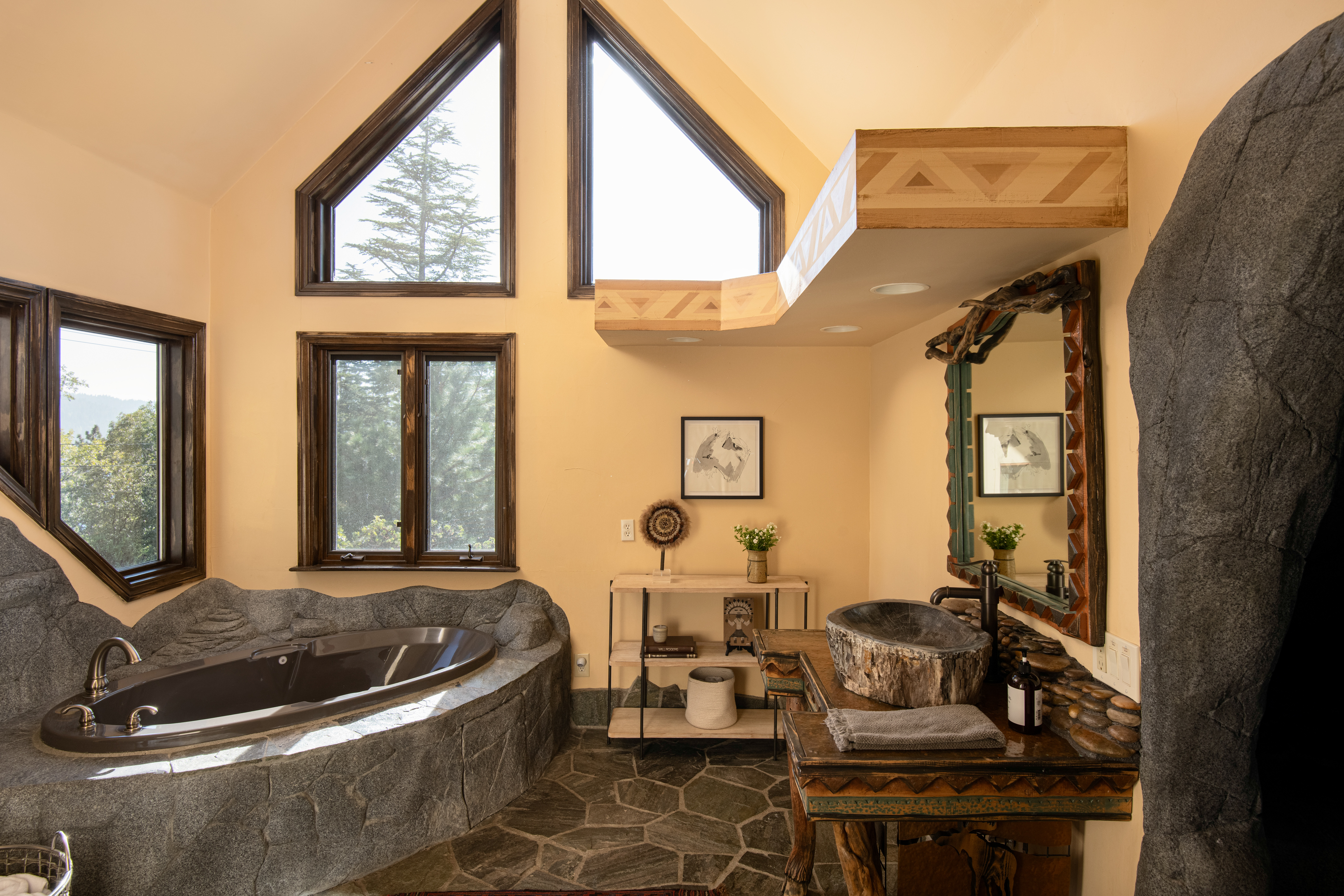 Spa-like bathroom with stone soaking tub, vaulted ceilings, and forest views