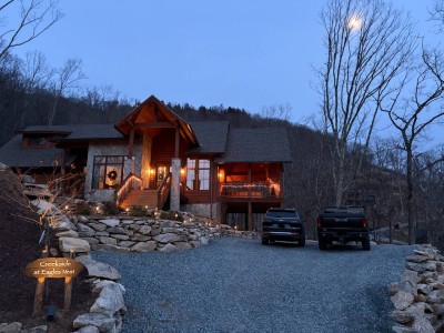 The cabin exterior shines in the evening with warm lighting, easy driveway access, and a welcoming atmosphere after a day exploring the mountains.
