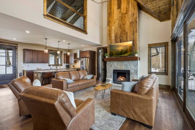 An open-concept great room with soaring ceilings connects the living area, kitchen, and dining space. Plush leather seating surrounds a stone fireplace, while large windows and sliding doors bring in natural light and easy access to the deck.