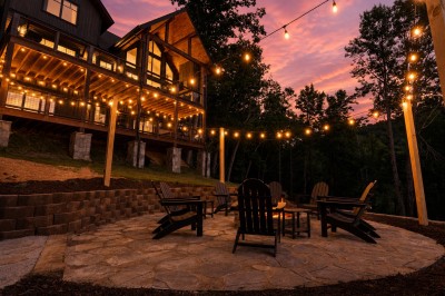 As the sky fades to twilight pink, gather around the fire pit on the stone patio beneath a canopy of string lights. Behind you, the cabin glows warm through every window — the perfect backdrop for a mountain evening well spent.