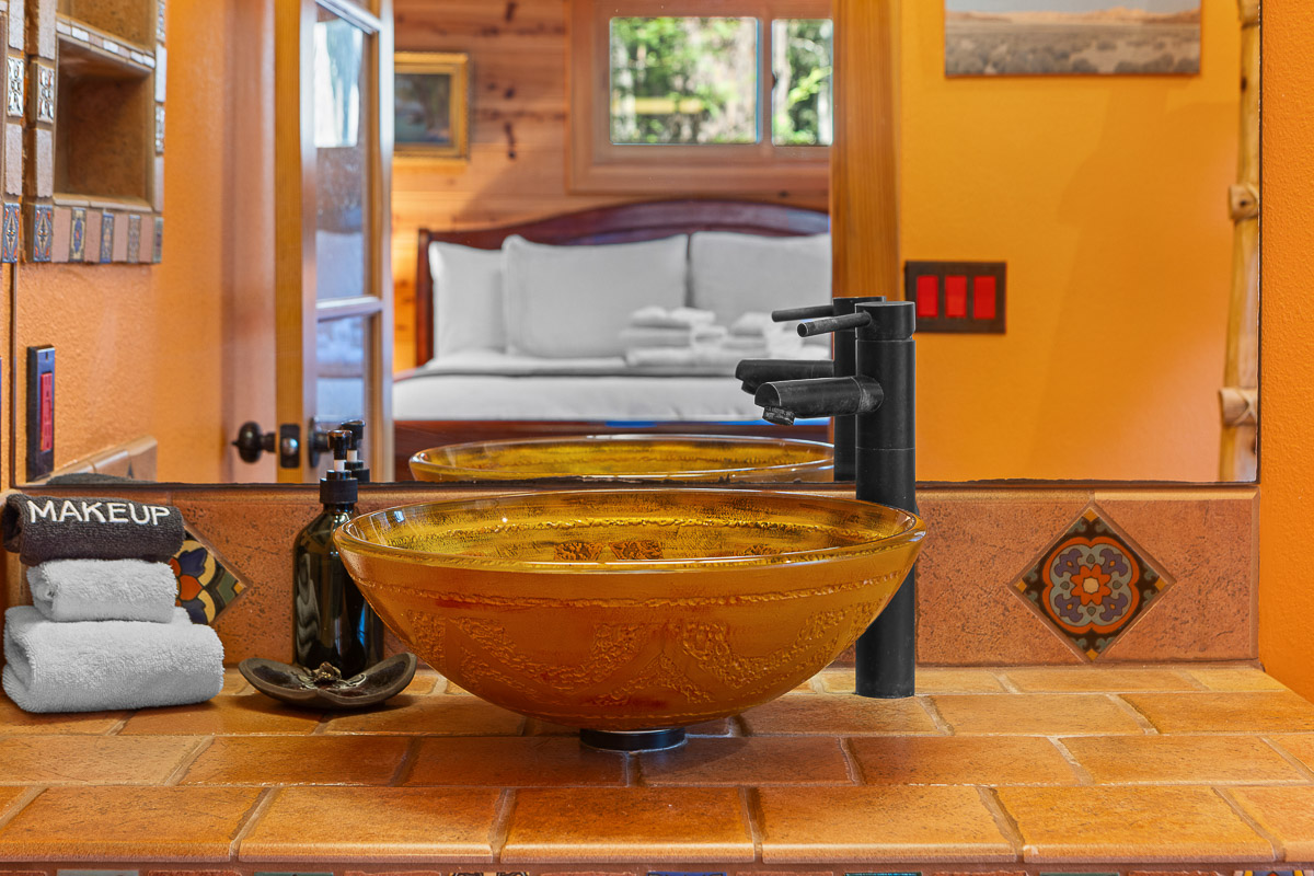 Warm rustic bath with amber vessel sink, view to plush cabin bedroom.