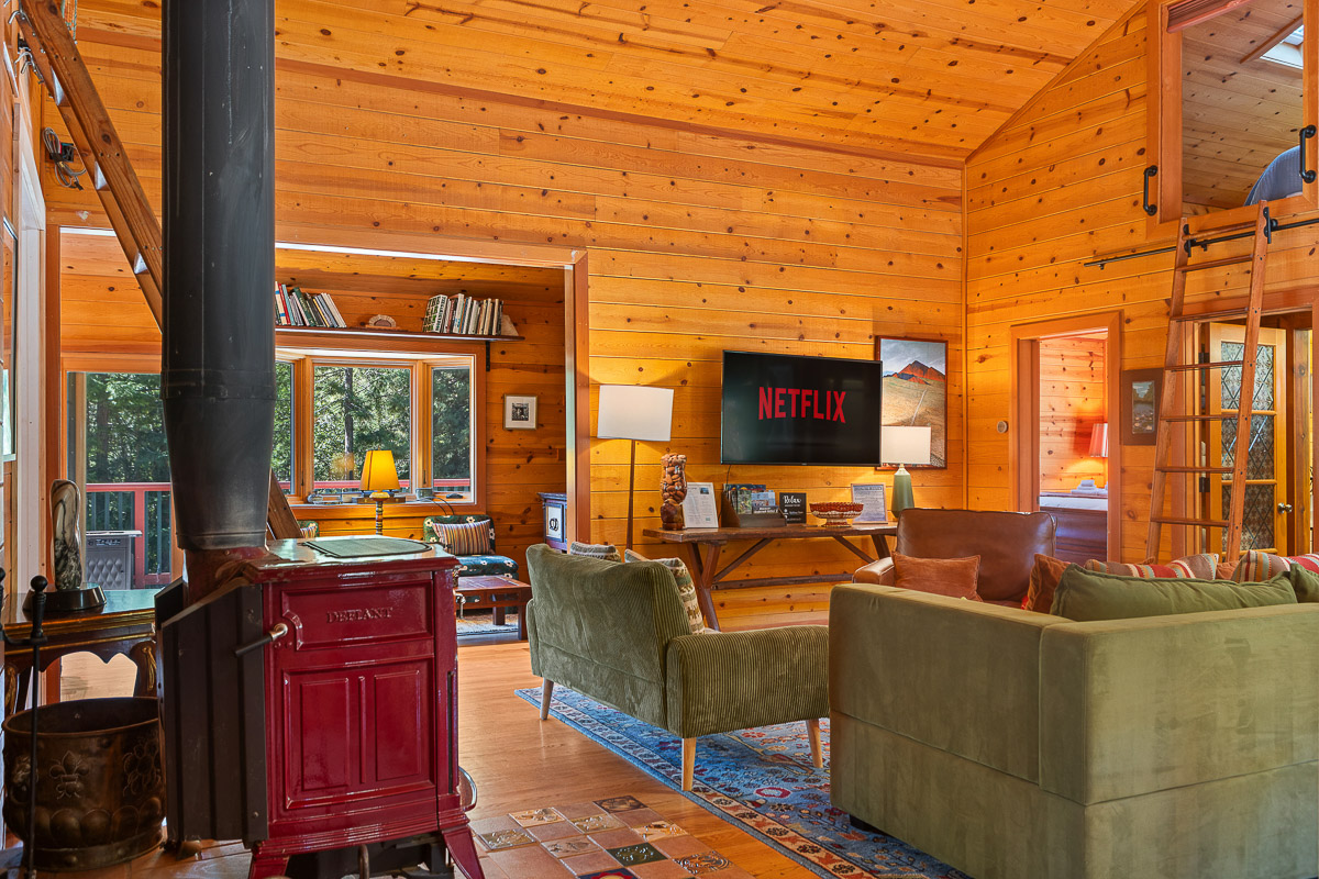 Cozy wood-lined lounge with stove, loft nook, smart TV, and forest views