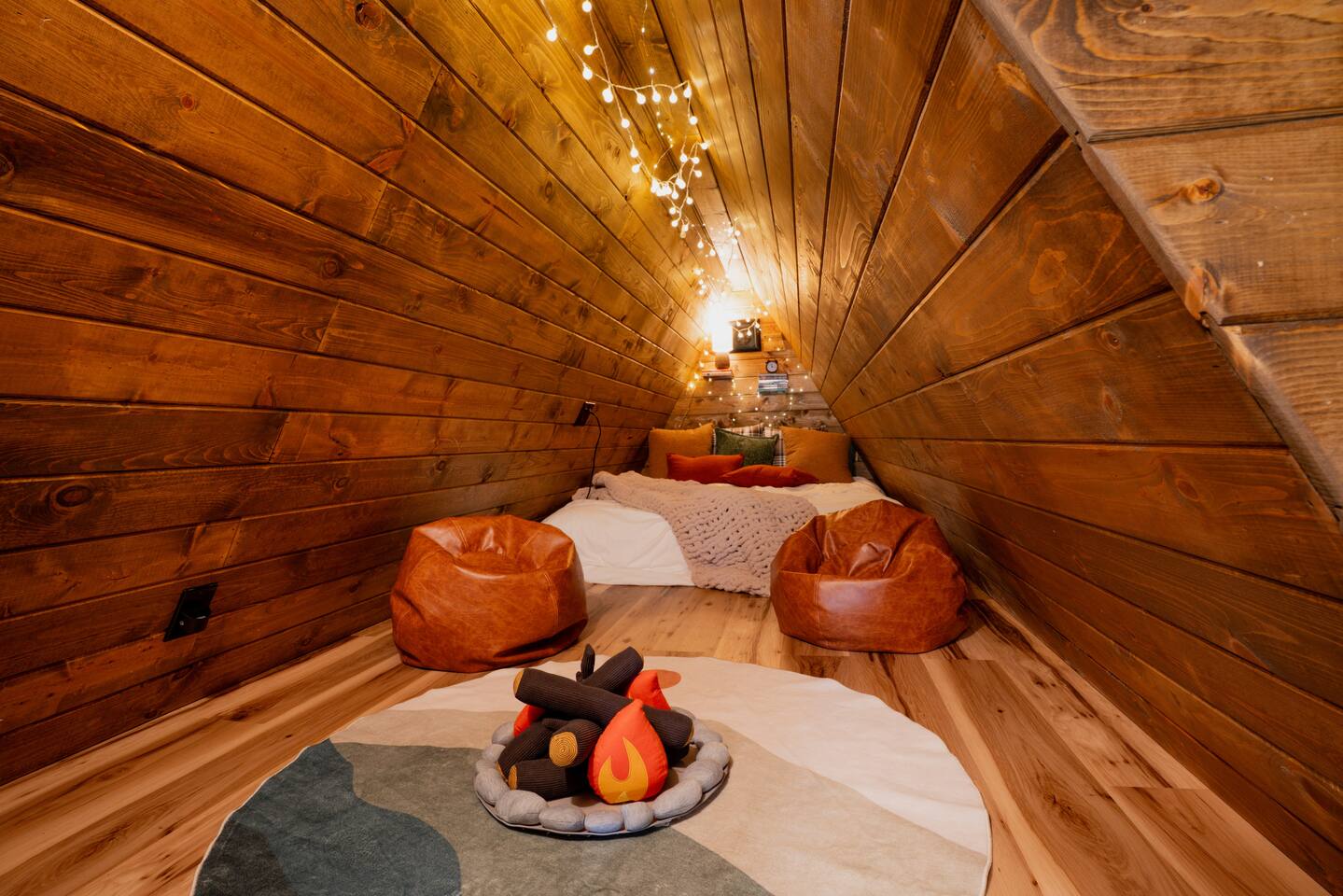 The finished secret attic bedroom, cedar-lined walls, string lights, bean bag chairs and a queen bed tucked under the eaves with a campfire rug on the floor.