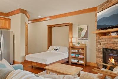 Board games and books line the shelf beside the built-in Murphy bed, which folds flush into custom cabinetry when not in use, opening up floor space near the kitchenette and the stone-corner fireplace glowing across the room.