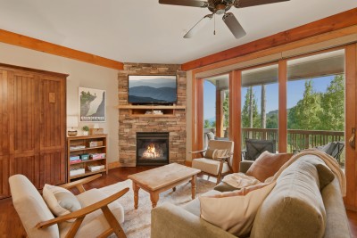 Gather around the stacked-stone gas fireplace beneath a mounted TV, with wood-framed armchairs and a deep sofa arranged for movie nights beside timber beams and hardwood floors, sliding doors opening onto the deck and its treetop mountain view.