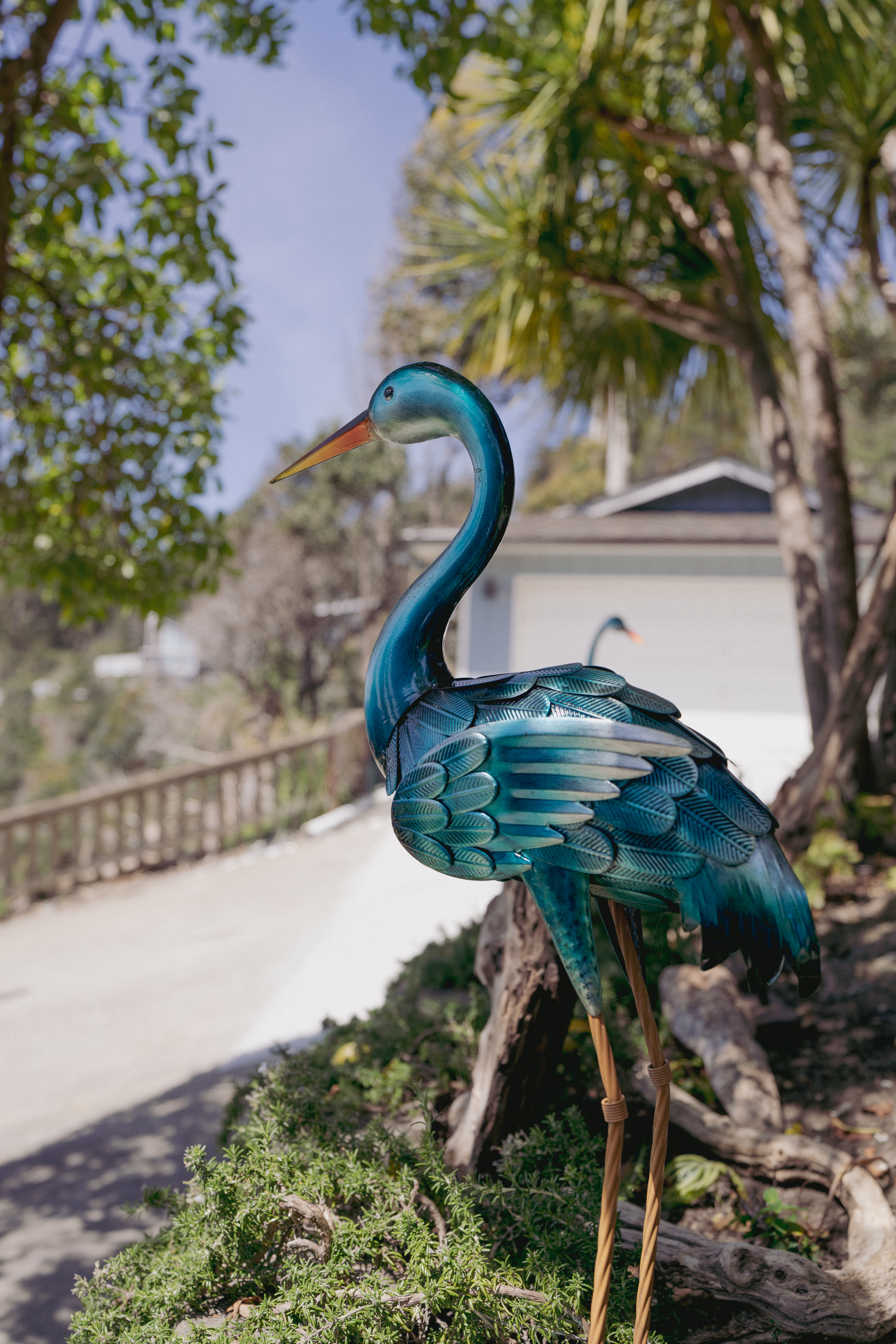 Sunny entry with teal heron sculpture and lush trees—welcoming charm