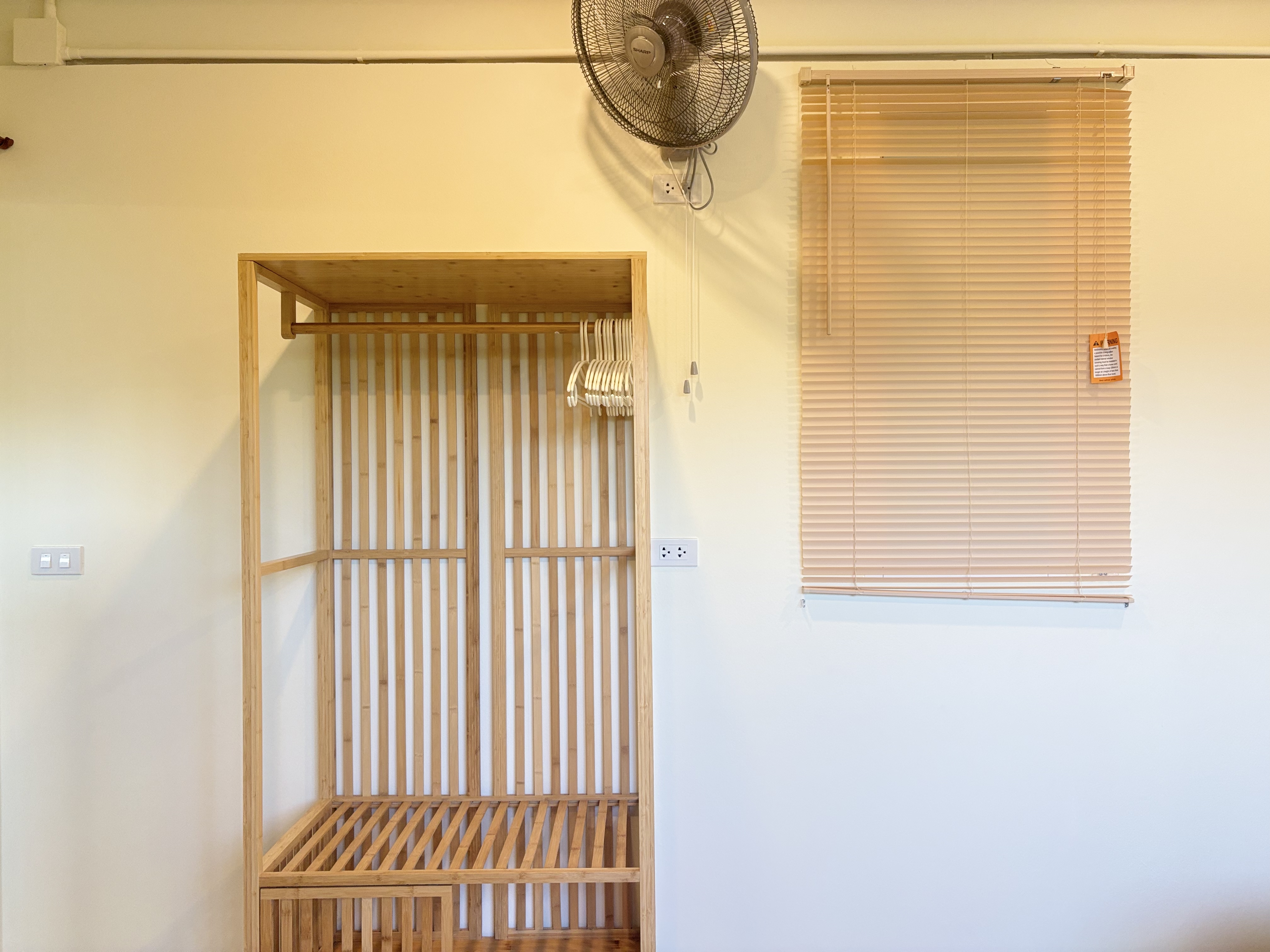 Minimalist room with open wooden wardrobe, window blinds, and cooling fan