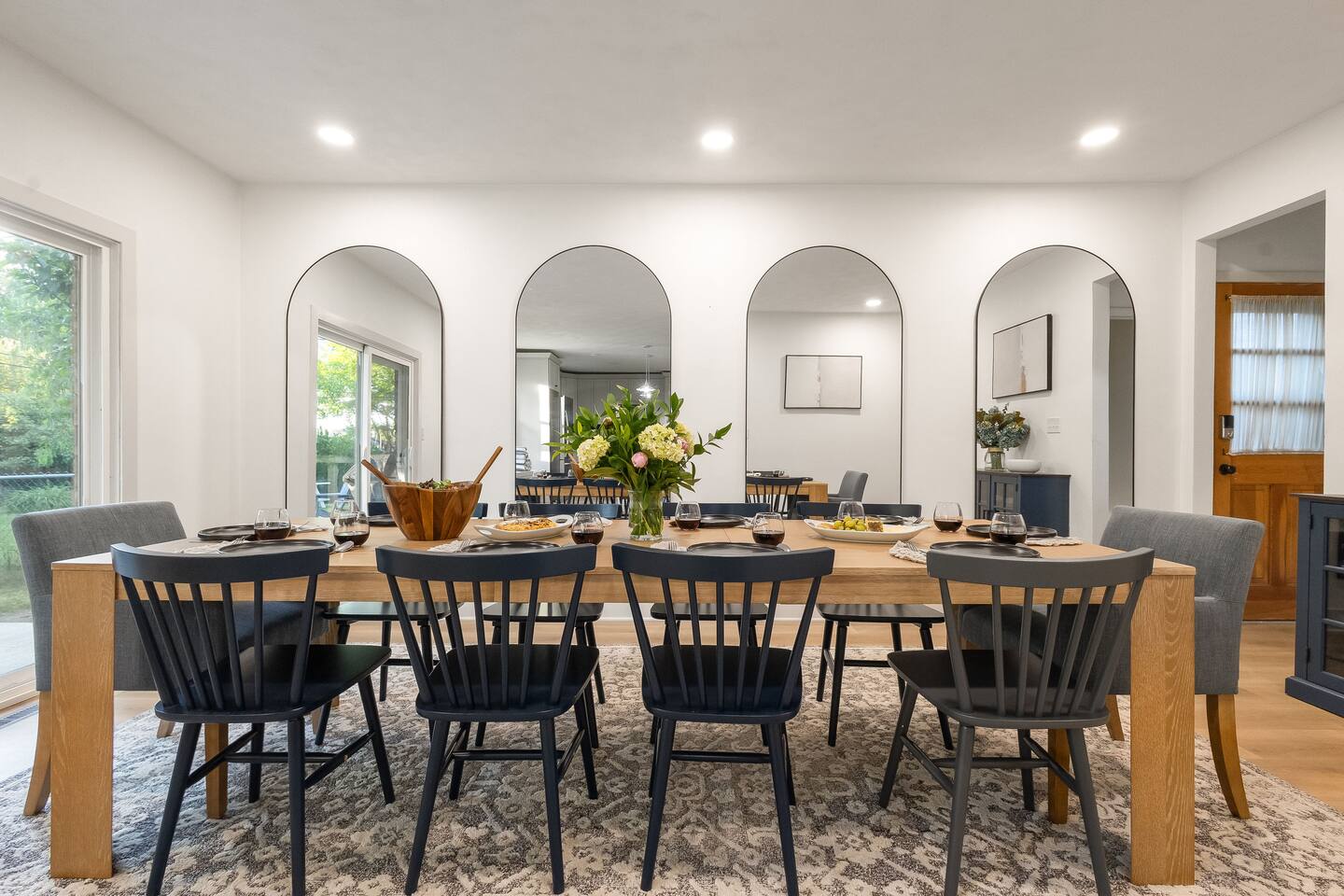 Long oak dining table set for ten under a wall of arched mirrors, with patio doors behind