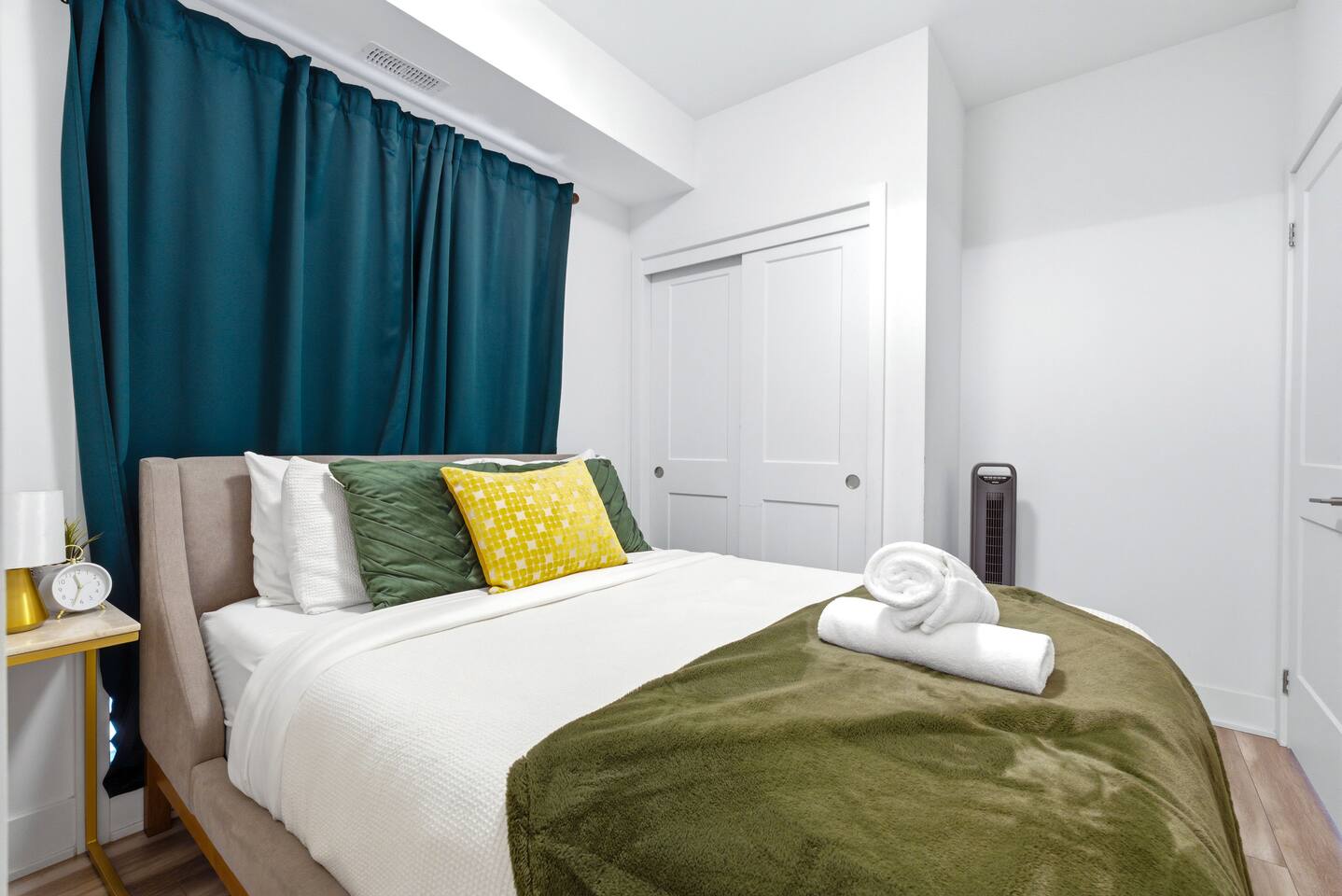Bright bedroom with crisp bedding, green accents, fresh towels, and dark curtains, creating a calm and comfortable space to rest.