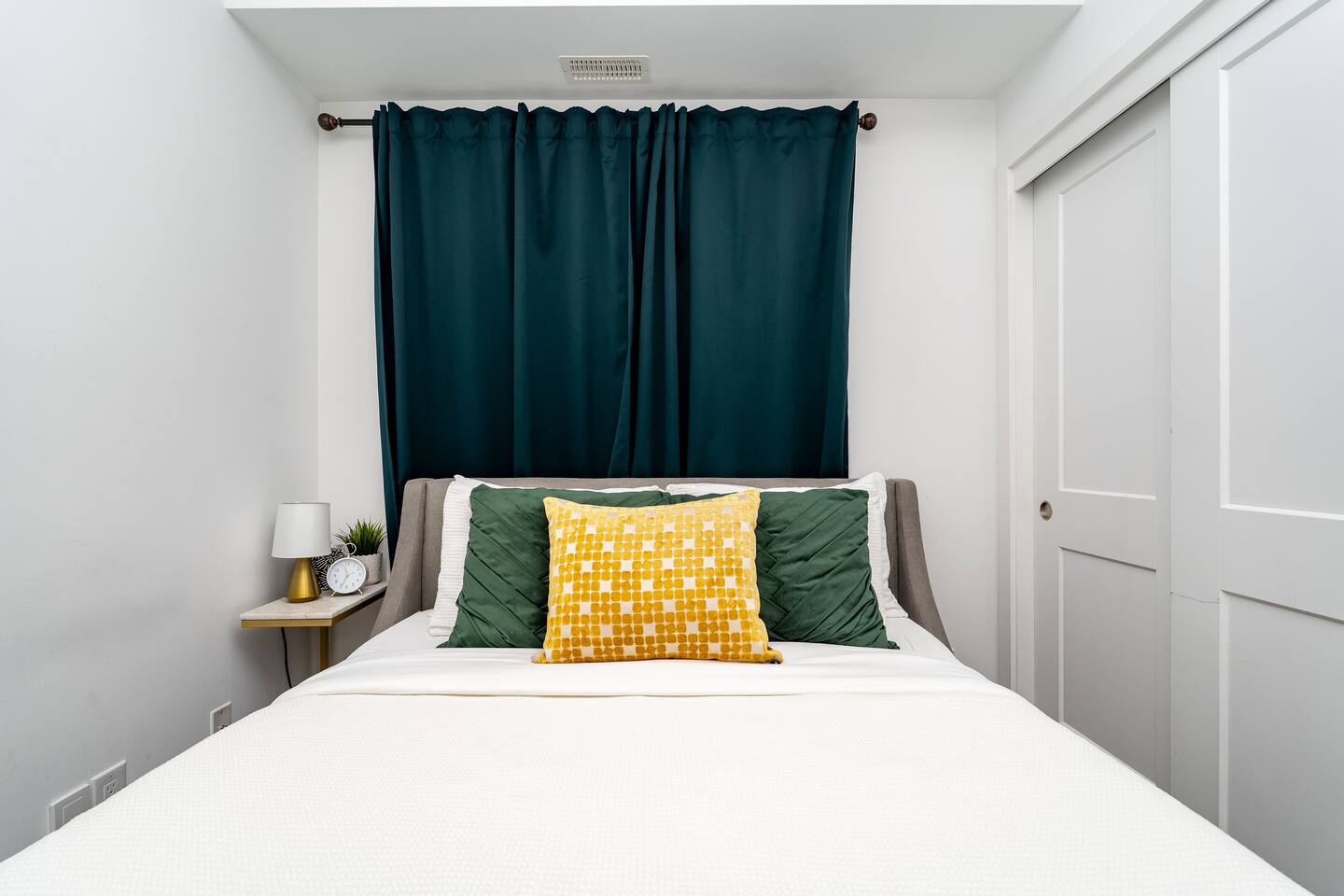 Bright bedroom with crisp bedding, layered pillows, and dark curtains, offering a simple and comfortable place to unwind.