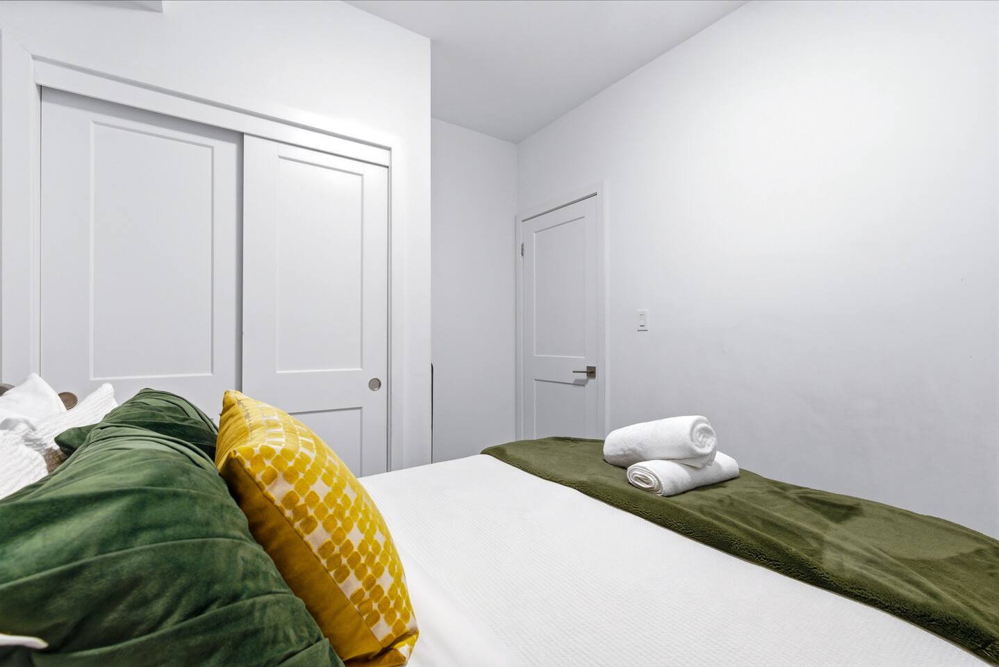 Clean bedroom with soft bedding, green and yellow accents, and fresh towels, offering a simple and restful space for guests.