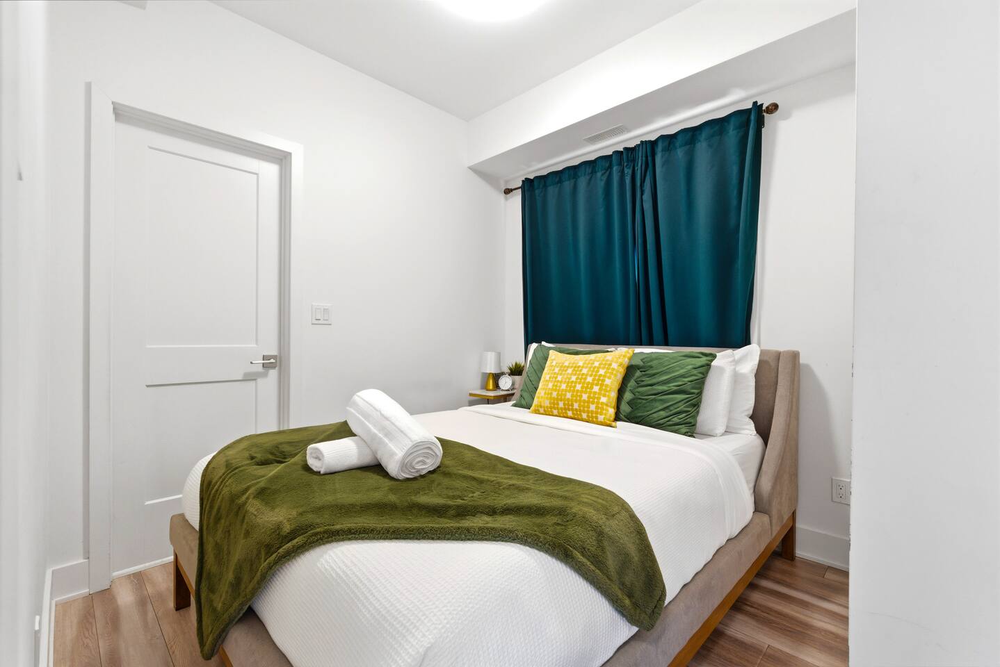 Cozy bedroom with crisp bedding, green and yellow accents, fresh towels, and dark curtains for a restful stay.