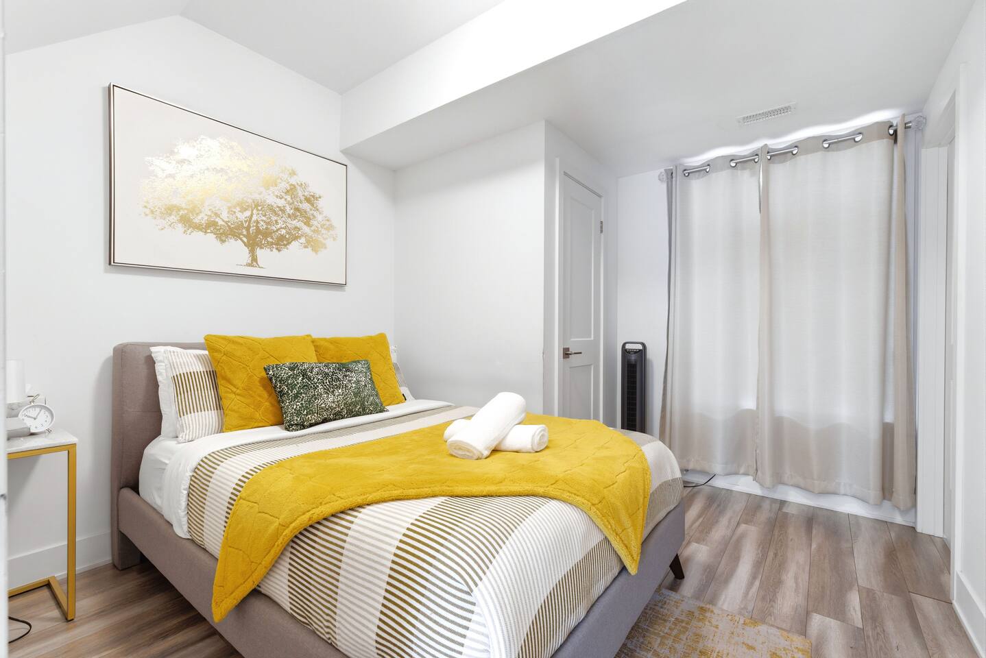 Bright bedroom with warm yellow accents, fresh towels, soft curtains, and wall art, creating a clean and cozy place to rest.