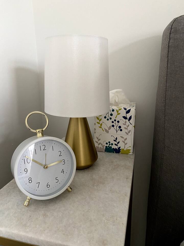Bedside table with a lamp, clock, and tissues, adding simple comfort and convenience beside the bed.