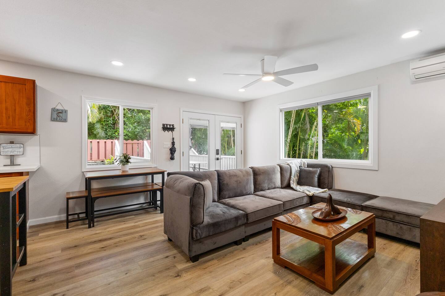 This sunlit living space features a plush sectional, warm wood accents, and lush views through large windows.