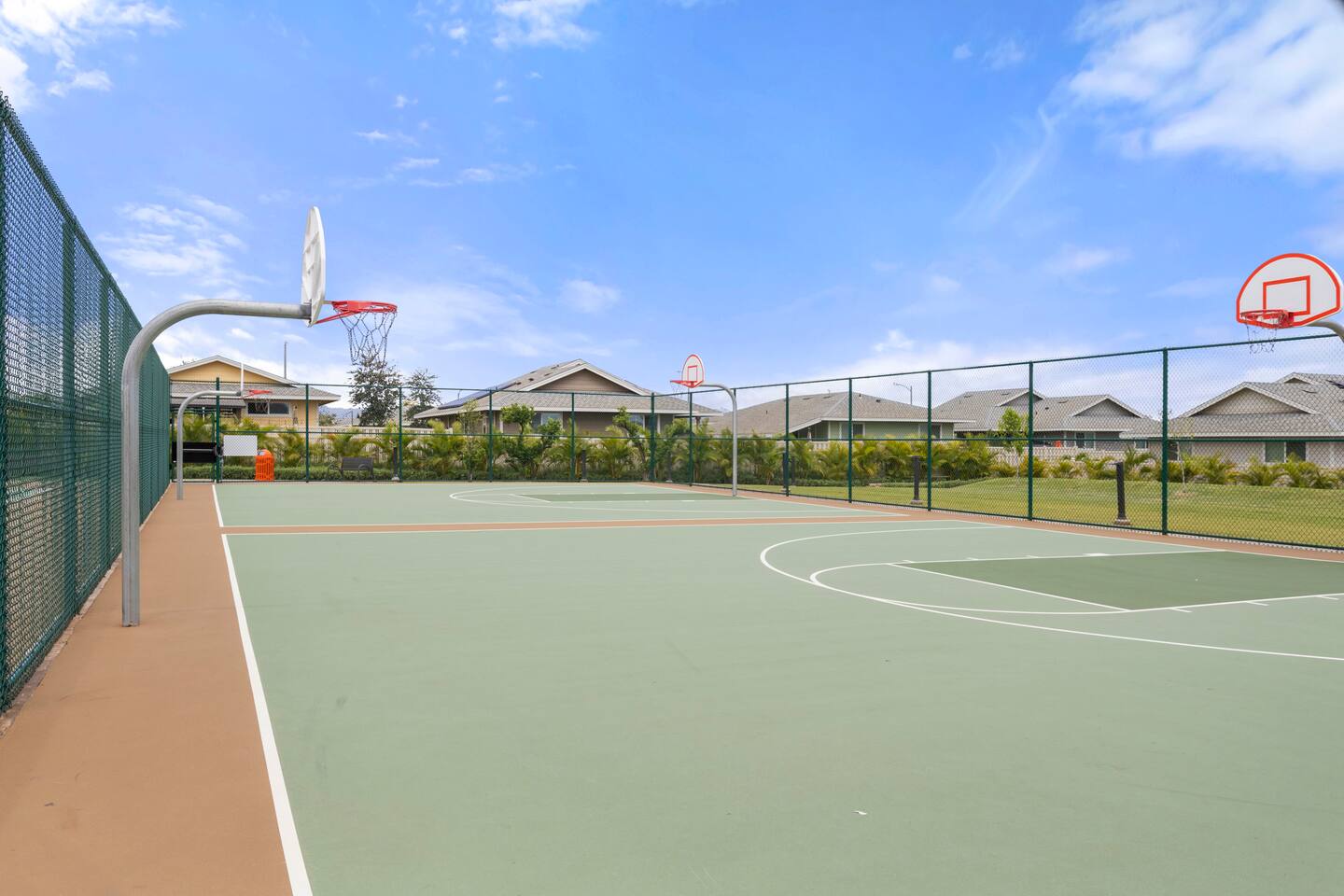 Outdoor basketball court with multiple hoops, marked lines, and residential backdrop behind chain-link fencing.