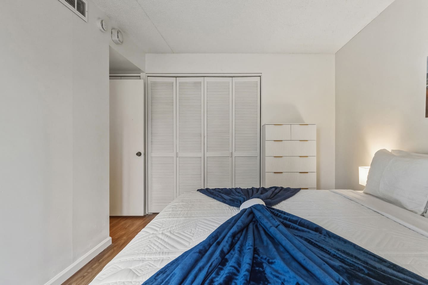 Rest easy in modern calm. White walls, wood floors, and a fan-folded blanket set the tone for quiet nights.