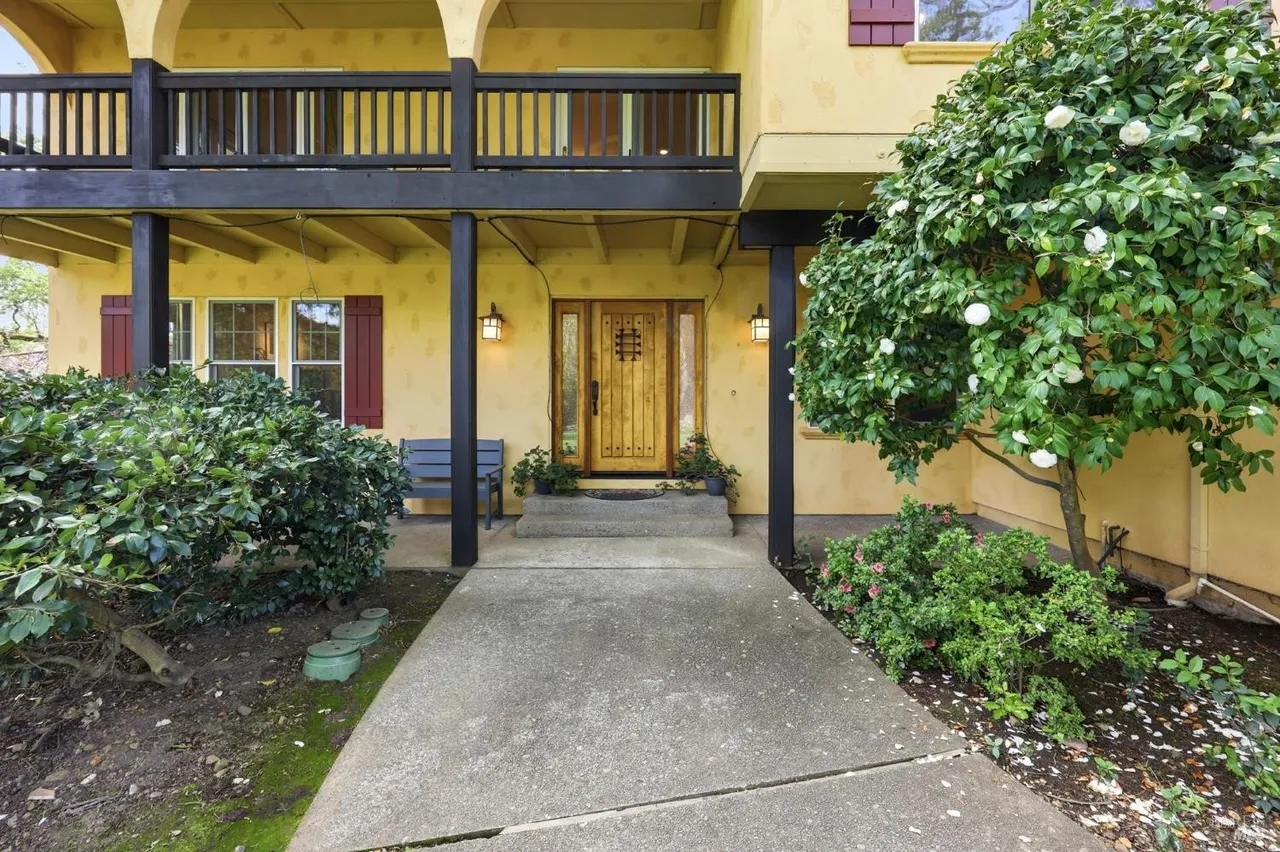 Welcoming entry with covered porch, lush landscaping, and charming balcony