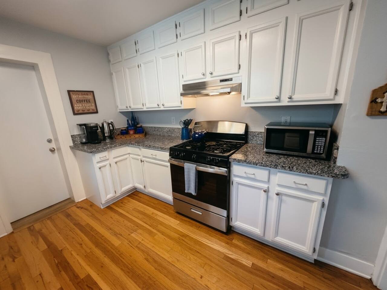 The full kitchen features bright white cabinetry, sleek granite countertops, stainless steel appliances, and rich hardwood floors. Fully equipped with modern essentials, it offers a warm, functional space for preparing meals.