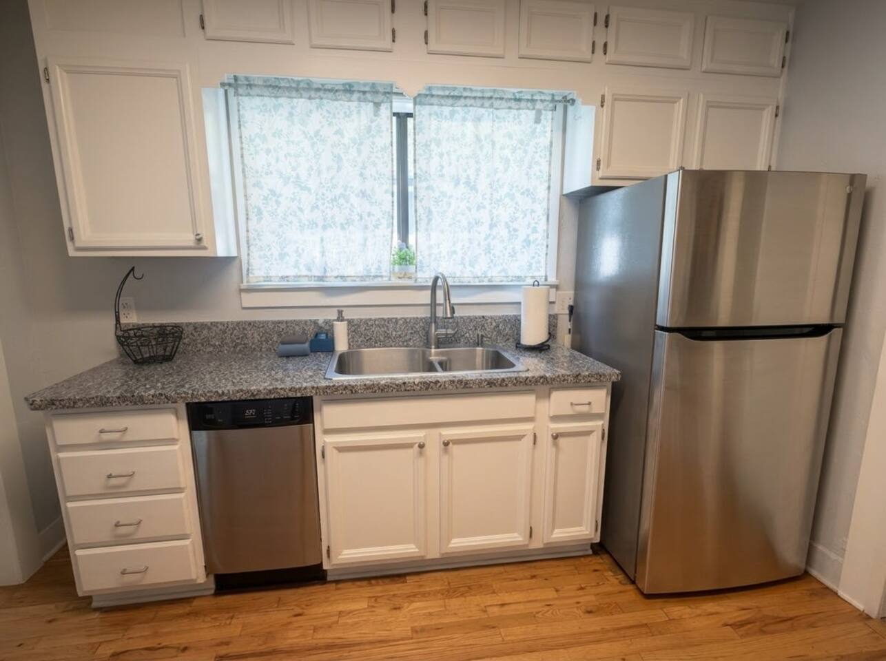 The full kitchen features bright white cabinetry, sleek granite countertops, stainless steel appliances, and rich hardwood floors. Fully equipped with modern essentials, it offers a warm, functional space for preparing meals.