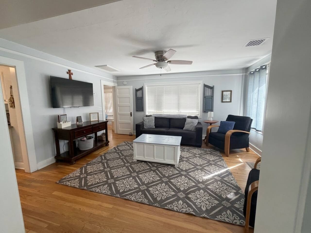 The living room at Virginia Blue features warm hardwood flooring, plush seating with a sleeper sofa, and a large wall-mounted flat-screen TV. Highlighted by natural light. The space offers a cozy and welcoming setting for guests to relax.