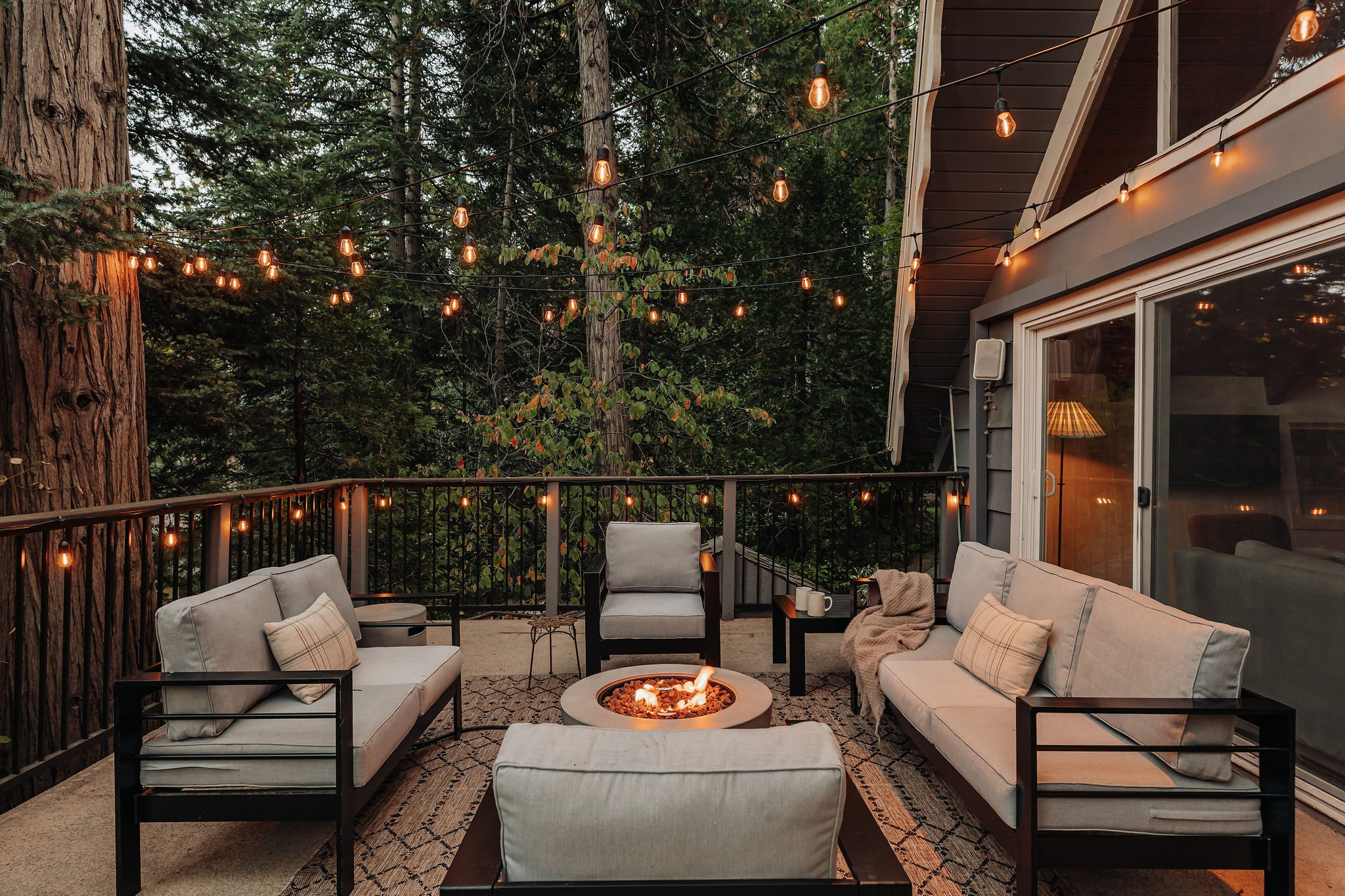 Forest-view deck with fire pit, lounge seating, and twinkling string lights