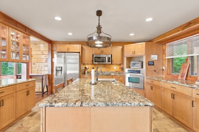 Kitchen with warm wood-toned cabinetry and flooring, granite countertops and a large center island, stainless steel appliances including a wall oven, cooktop, and refrigerator, plus glass-front upper cabinets and a hanging wrought-iron pendant light.