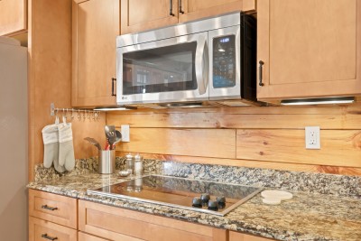 A wide view of a kitchen with granite countertops, stainless steel refrigerator, microwave, and dishwasher, rustic wood cabinetry, and an open sightline through to a great room with tall peeled log posts and a wall of large windows in the background.