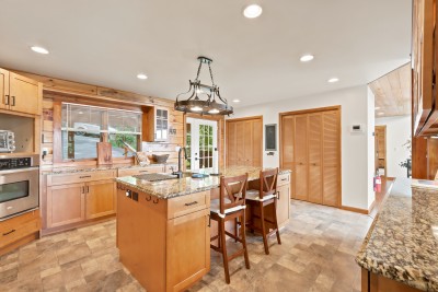 Kitchen with a granite island lined by several wooden bar stools for casual seating, granite countertops, glass-front cabinets, stainless appliances, and a clear sightline through to the great room's wall of windows and layered mountain views beyond.