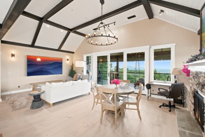 One room does it all: sofa, dining table, fireplace, and a wall of windows aimed straight at the ridgeline. Vaulted ceilings and beams give it real mountain-lodge scale.