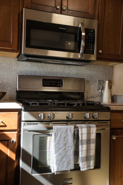 Stylish kitchen with a stainless gas range beneath a black shiplap hood, a six-seat quartz island, double ovens, and a farmhouse sink, vaulted and stone-backed, built for cooking as a group.