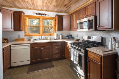 This fully equipped kitchen centers on a vaulted, stone-backed space with a six-seat quartz island, gas range, double ovens, and a farmhouse sink beneath a sunlit window, warm, functional, and built for cooking together.