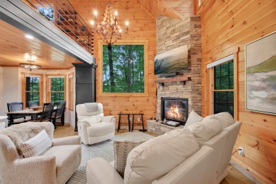 The great room opens straight into the dining area, with a stacked stone hearth at the center and windows full of green on both sides.
